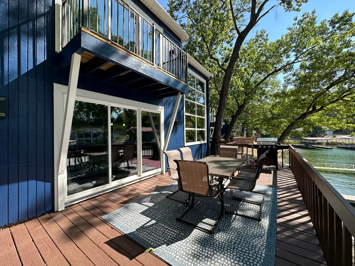 The image shows the condo's exterior with expansive windows overlooking the peaceful cove, a spacious deck area set for outdoor dining, surrounded by lush trees. The scene is bathed in soft sunset light, inviting relaxation and cheerful gatherings with friends and family.
