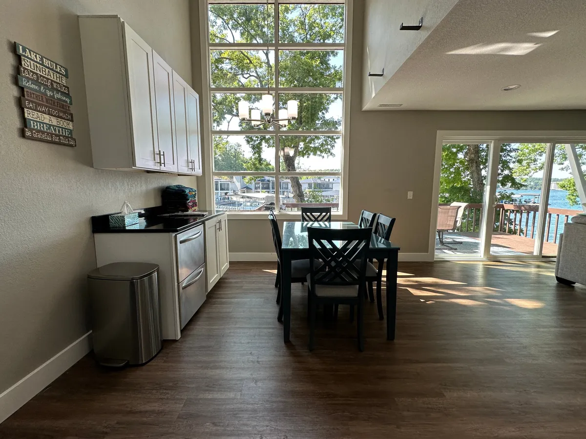 This image shows a bright, airy dining area with a large table positioned next to expansive windows that let in abundant natural light. The background includes a modern kitchen with sleek appliances and countertops, emphasizing the openness and flow between the spaces. The scene evokes a sense of communal gathering, with views that hint at the lakeside location.