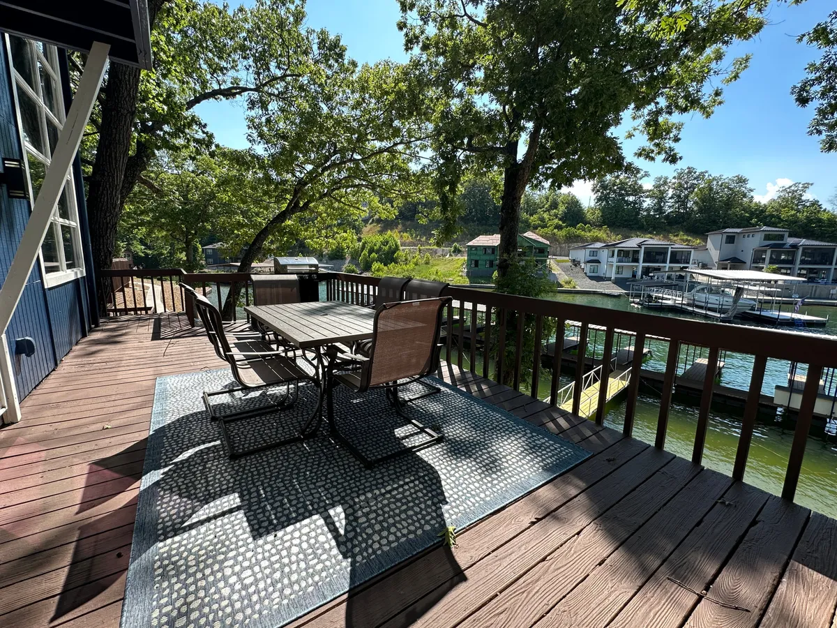 An inviting outdoor dining table set under the trees with a direct view of the sparkling water. The setup suggests space for morning coffee, meals, or casual gatherings while enjoying the natural surroundings. The table area is close to the water, emphasizing easy access to dockside leisure.