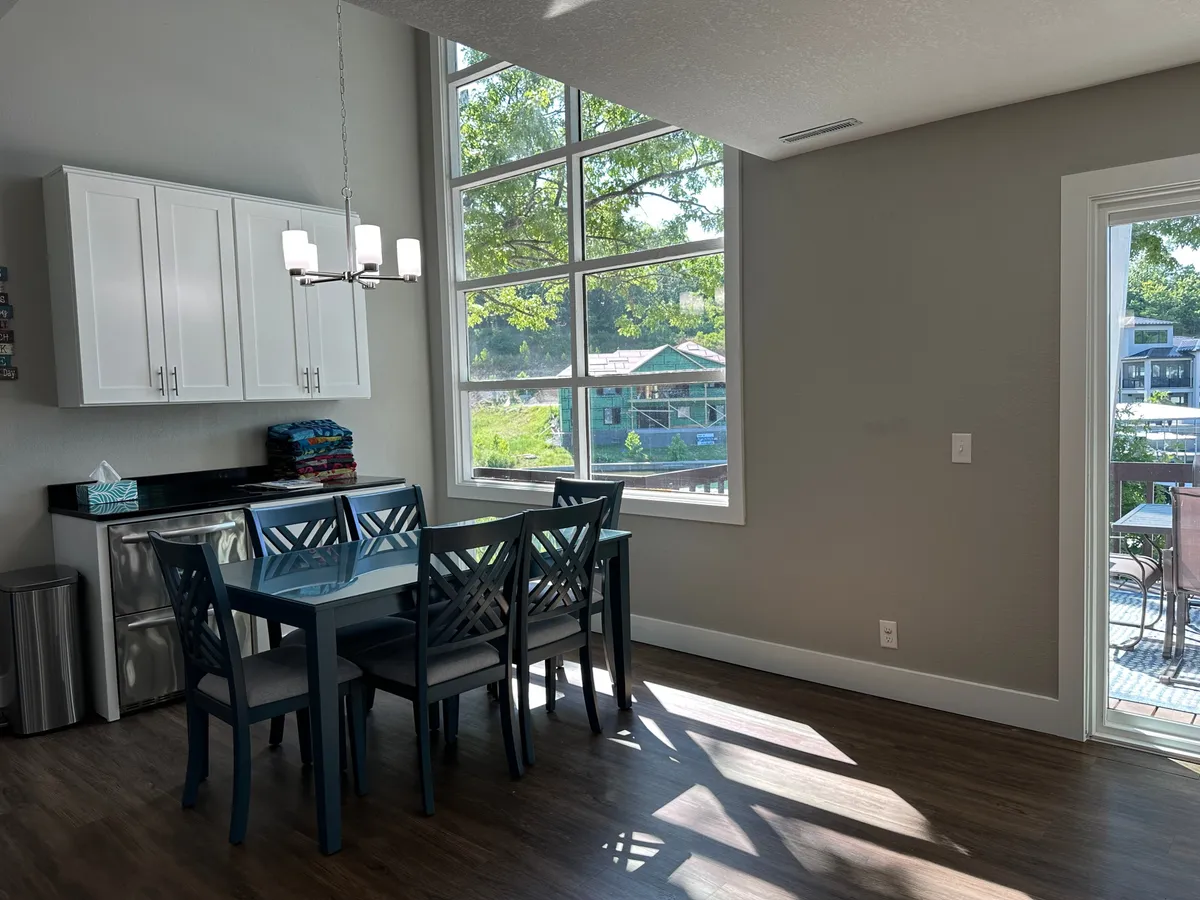 A warm, welcoming dining area with a wooden dining table surrounded by chairs, set near large windows that let in ample sunlight. The kitchen cabinetry and open space suggest a sociable environment ideal for group meals and festive gatherings.