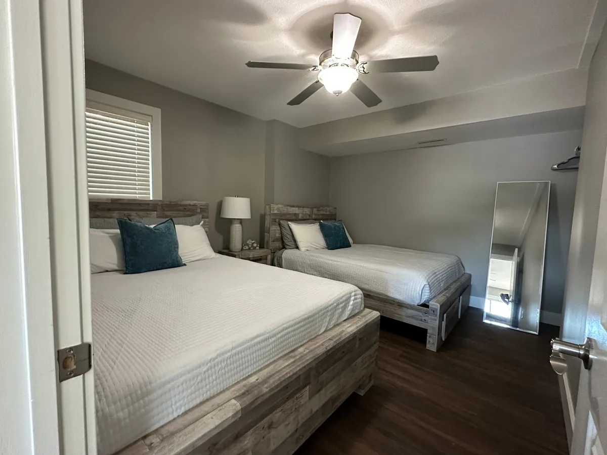 A cozy bedroom featuring two beds with wooden frames and plush bedding, a ceiling fan with light, a bedside table with a lamp, and a large mirror. The room feels relaxing and comfortable, making it suitable for families or groups with flexible sleeping arrangements.
