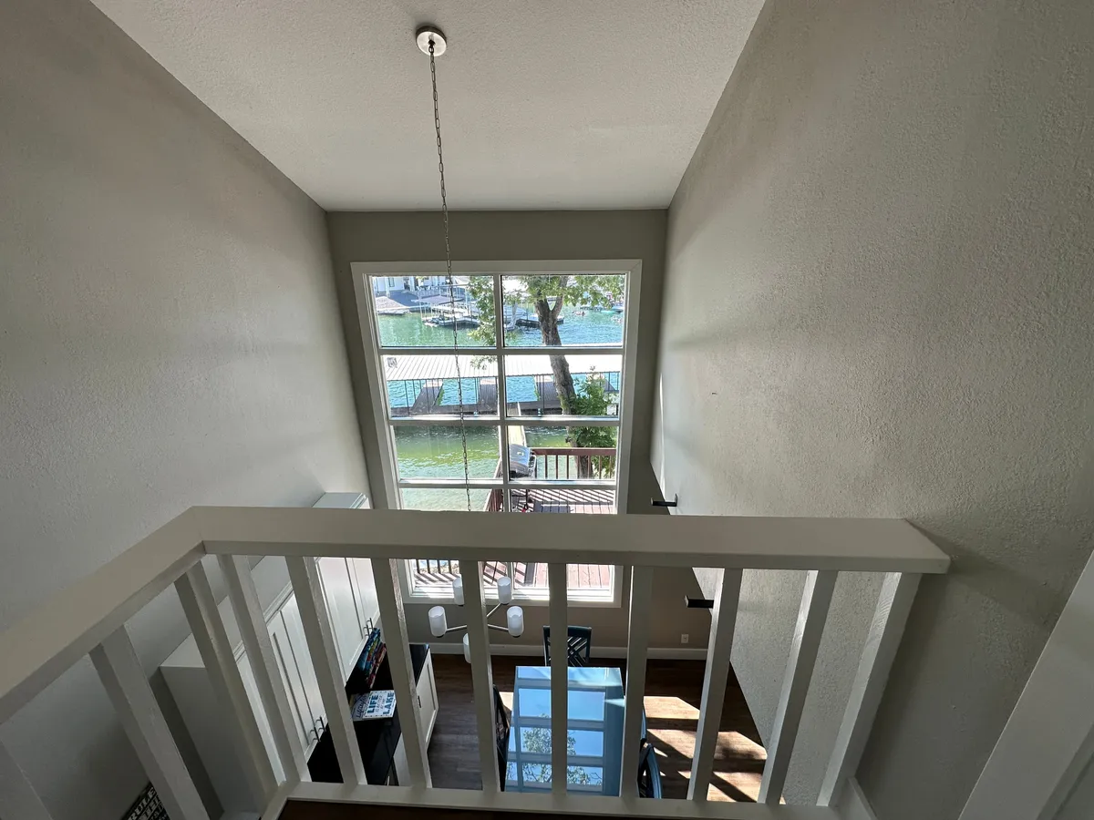 A bright interior scene featuring large windows that open up to reveal a beautiful water view. A spacious dining table sits beside the windows, and just beyond is a balcony area overlooking the lake. The scene is set for casual dining, socializing, and relaxation with direct visual access to the outdoors.