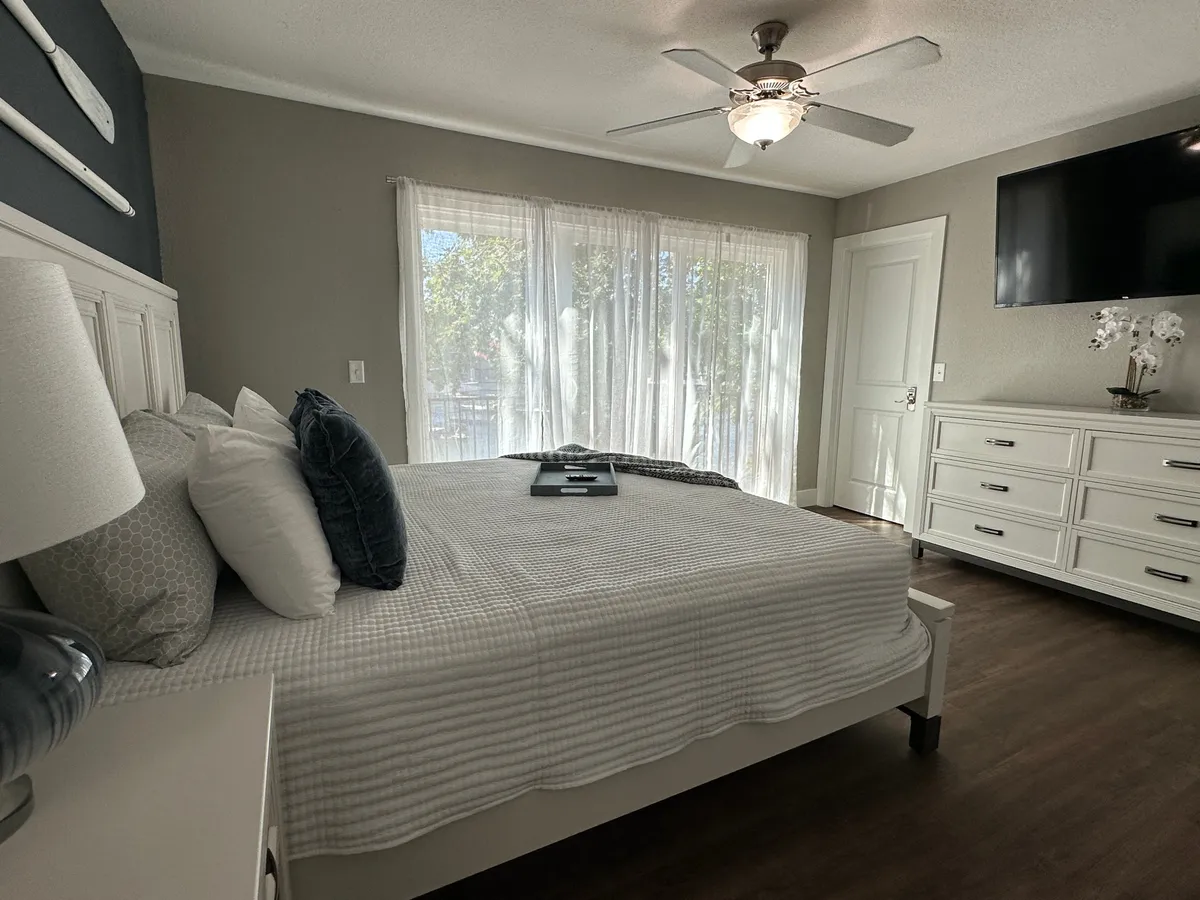 A serene bedroom with a bed featuring plush pillows, large windows draped with sheer curtains that let in natural light, a TV mounted on the wall, and a dresser topped with a decorative plant. The atmosphere is tranquil and inviting, perfect for relaxation and unwinding in private comfort.