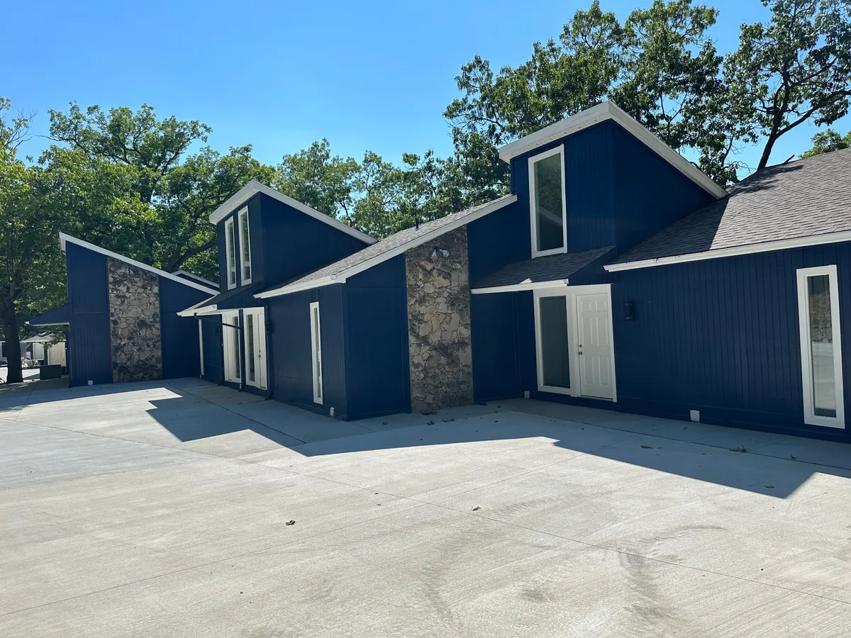 The image showcases the inviting exterior of a modern lakeside property, with blue walls, stone accents, and angled roofs nestled among natural surroundings. The ambiance is both contemporary and serene, suggesting comfort and elegance. The patio area encourages family gatherings and outdoor meals in a beautiful setting.